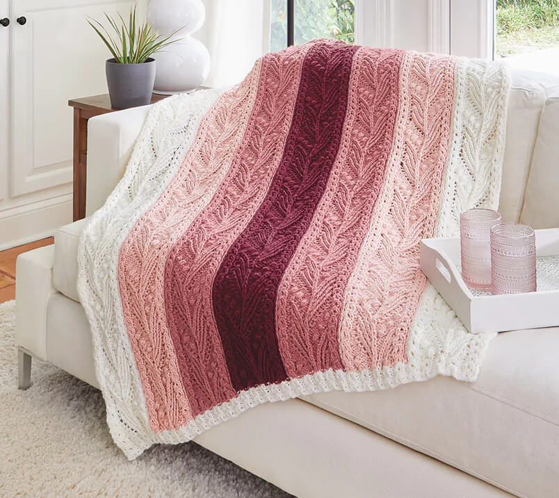 A knitted afghan made of 7 white and pink strips of knitting. Each strip has a leaf-like pattern to the knitting.